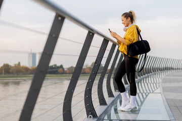 Cheerful woman in a yellow hoodie enjoys a sunny afternoon stroll along a modern riverside promenade © BGStock72