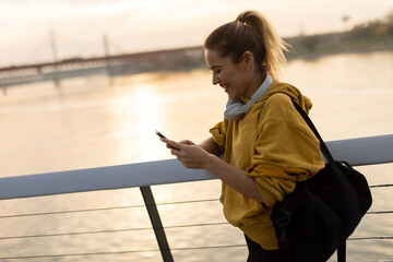 Obraz premium A woman in a yellow hoodie enjoys a peaceful moment by the river while checking her phone during sunset in a serene urban setting