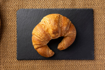 A freshly baked, golden brown croissant rests atop a textured black slate surface, contrasting the...