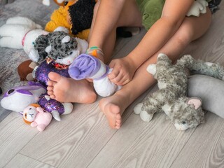 Close-up of the bare feet of a little girl sitting on the floor among a pile of soft toys, holding...