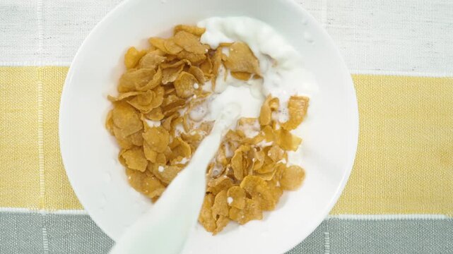 Close up of a white bowl of cereal with milk being poured into it. Macrography of white milk pouring in to bowl with yellow cornflake and splashing milk with isolated white background. Pabulum.