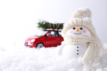 Cute decorative snowman and toy car with fir tree branches on artificial snow against light background, selective focus