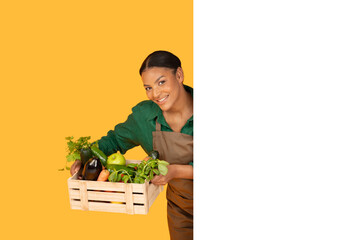 A smiling black woman in an apron holds a crate filled with various fresh vegetables, showcasing her passion for healthy eating and agricultural produce, white blank board for ad