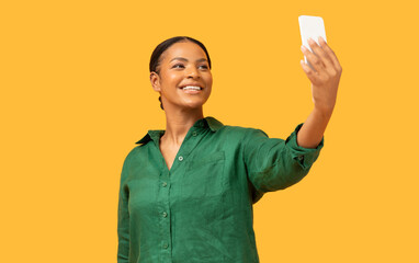 A black woman wearing a green shirt smiles broadly while holding her smartphone to take a selfie. The bright yellow background adds a vibrant touch to the moment.