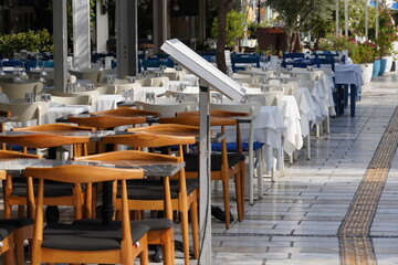 An empty outdoor restaurant features a menu stand in the foreground, inviting passersby to browse. The tables, adorned in blue and white, recede into the background in a diminishing perspective.