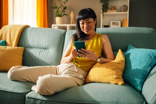 Asian woman enjoying her smartphone on green couch, colorful living room - Powered by Adobe
