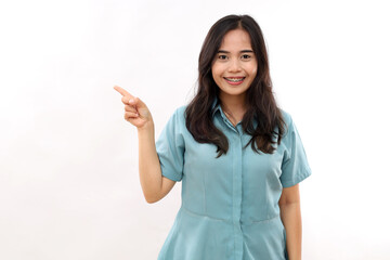 Beautiful Asian woman indicates aside, shows promo, gives advice, makes choice, smiles pleasantly, wears casual shirt, stands against white studio wall, tells about product details