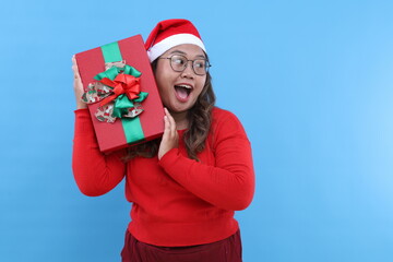 Christmas presents delivery. Cheerful young Asian woman in Santa hat holds Xmas gift, posing with red wrapped box with ribbon bow blue studio background. Holiday cheer and kindness concept