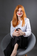 Portrait of a red-haired girl in a white shirt. Woman sitting on a chair in the studio against a gray background.