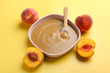 Delicious baby food with spoon in bowl and fresh peaches on yellow table