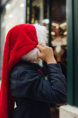 Little school kid boy in Santa hat on Christmas market. Funny happy child in fashion winter clothes making window shopping decorated with gifts. Holidays, Christmas, childhood and people concept