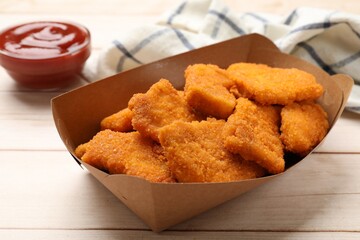 Tasty chicken nuggets in paper container and sauce on wooden table, closeup