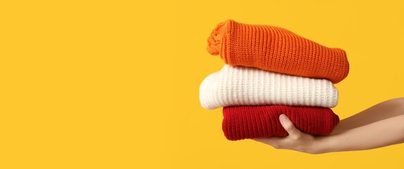 Female hands holding stack of warm clothes on yellow background with space for text