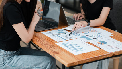 Two women discussing financial data with laptops and printed charts, showcasing teamwork and...