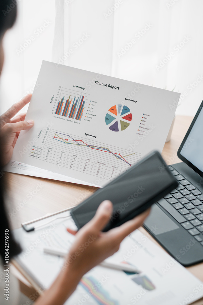 Poster close-up of a person reviewing a financial report with graphs and charts, using a smartphone and lap