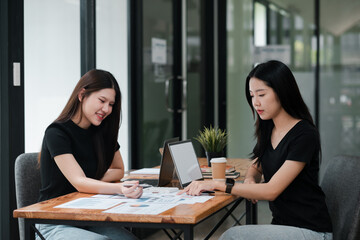 Two women working together in a contemporary office setting, discussing business plans with laptops and documents.