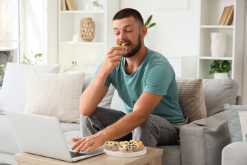Young man with laptop eating tasty muffin at home
