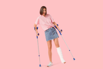 Young woman with broken leg and crutches on pink background