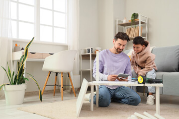 Father with tablet computer and his little son assembling shelf unit at home