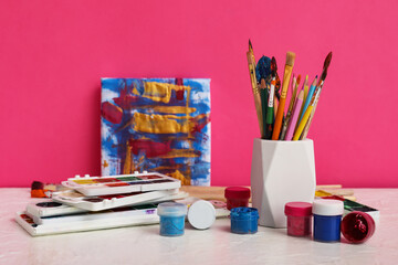 Artist brushes, paints and painting on white table against pink background