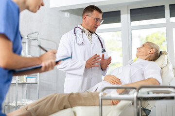 Obraz premium Male beautician, together with a nurse, examines the face of an elderly woman and plans facelift procedures.