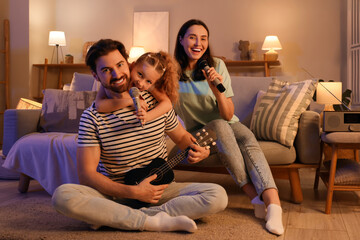 Happy family with microphones and ukulele singing karaoke on sofa at home in evening