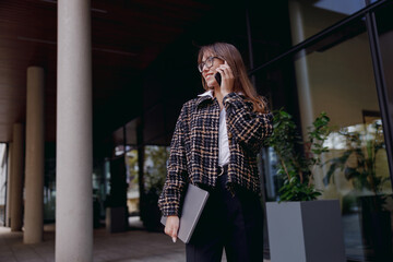 A confident woman in a trendy plaid jacket chats happily on her mobile phone in a modern business setting