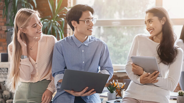 Cheerful young multiracial coworkers sharing business ideas, using digital tablet and laptop, office interior