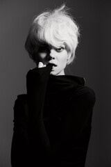 Elegant woman with short blonde hair, wearing a black turtleneck, looking thoughtfully at the camera. Dark background enhances the striking contrast.