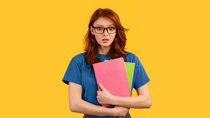 End-of-semester exams. Shocked student girl in eyeglasses holding books standing on yellow studio...