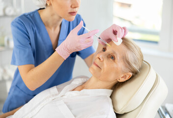 Elderly woman undergoing botulinum therapy session at aesthetic medicine clinic. Confident cosmetologist making injections between eyebrows of patient with insulin syringe to correct facial wrinkles