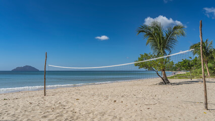 The beach volleyball net is stretched on poles. Footprints in the sand. The waves of the turquoise ocean are foaming. Green tropical vegetation on the shore.The silhouette of the island on the horizon