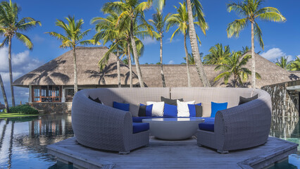 A place of rest at the resort. A semicircular wicker sofa on a board platform in the swimming pool. There are cushions on the soft seats. A round table in the center. Building with a thatched roof.