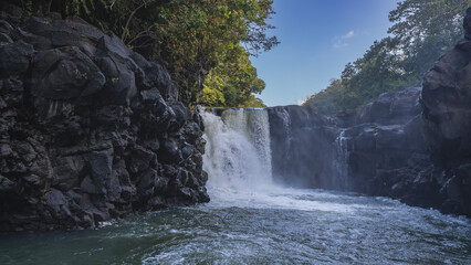 A beautiful tropical waterfall. Streams flow down from the cliff ledge into the river. Splashes, foam. Green vegetation on the rocks. The blue sky. Mauritius. Grand River South East waterfall 