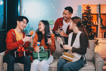 Group of young Asian man and women as friends having fun at a New Year's celebration, holding gift boxes standing by Christmas tree decoration, midnight countdown Party at home with holiday season.