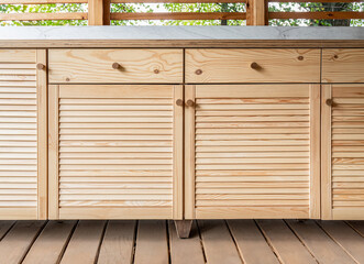 Wooden cabinets of outdoor kitchen with louvered doors, drawers, wooden handles and plywood countertop, stand on a wooden terrace