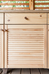 Wooden cabinets of outdoor kitchen with louvered doors, drawers, wooden handles and a white plywood countertop, stand on a wooden terrace close-up view