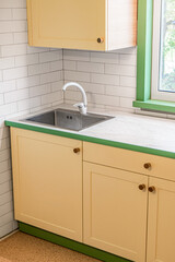 Fragment of kitchen with yellow cabinets with wooden handles features a plastic countertop with a marble-like finish, and upper cabinets installed around the window