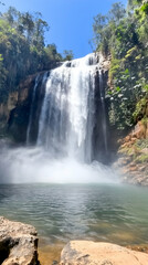 Fototapeta premium A cascading waterfall flows into a pool of water, surrounded by lush green foliage.