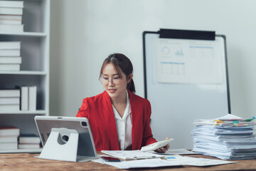 Focused and Ready: A confident businesswoman in a red blazer sits at her desk, diligently reviewing paperwork and utilizing a tablet in a well-organized and professional workspace.  