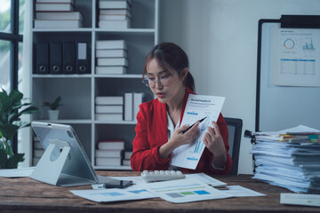 Focused Businesswoman Reviewing Data: A detail-oriented businesswoman in a red blazer sits at her...