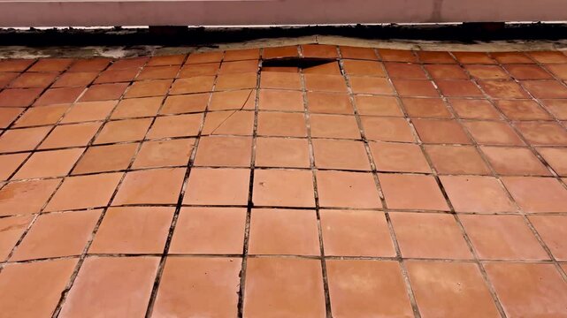 A distressed and uneven outdoor tile floor, showing significant damage and dislodged tiles. The image captures the effects of weathering and wear, emphasizing structural deterioration and maintenance 