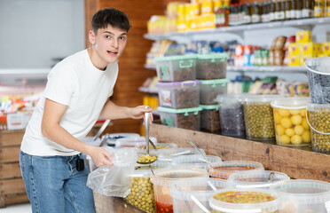 Positive young guy making routine purchases in local grocery store, scooping marinated olives from plastic buckets and placing in poly bag
