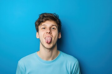 Fototapeta premium Portrait of a young caucasian man sticking his tongue out isolated on blue background