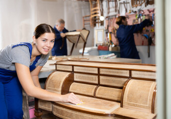 Skilled female furniture repair specialist in uniform restoring Victorian dresser during workday in workshop