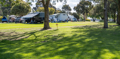 Close-up background texture of a grassy, unpowered campground under the shade of trees in a caravan park, with a blurred view of RVs and campers at the campsites in the distance.