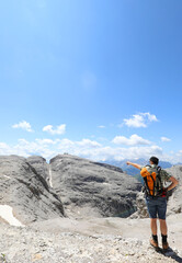 hiker with a backpack points at the view of the Sasso Pordoi mountain range in the Dolomites mountains in the European Alps in northern Italy