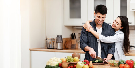 Happy Loving Family Concept. Young man cooking salad at kitchen, woman embracing him, copy space