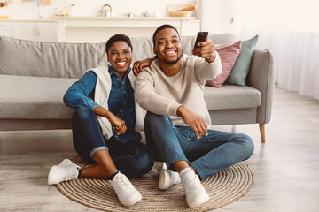 Leisure And Entertainment. Smiling African American couple watching TV show or film, guy holding remote control. Young man and woman enjoying free time sitting on floor carpet at home in living room