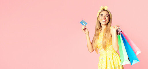 Shopping Online. Excited beautiful blonde girl holding credit card and paper bags over pink studio wall, copyspace